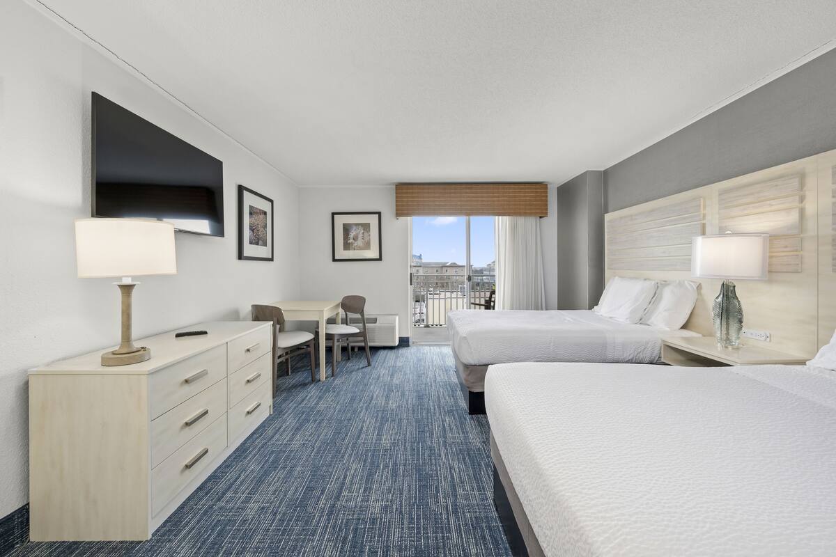 A hotel room with two beds, a desk, a chair, a dresser, and a large window with a view of buildings.