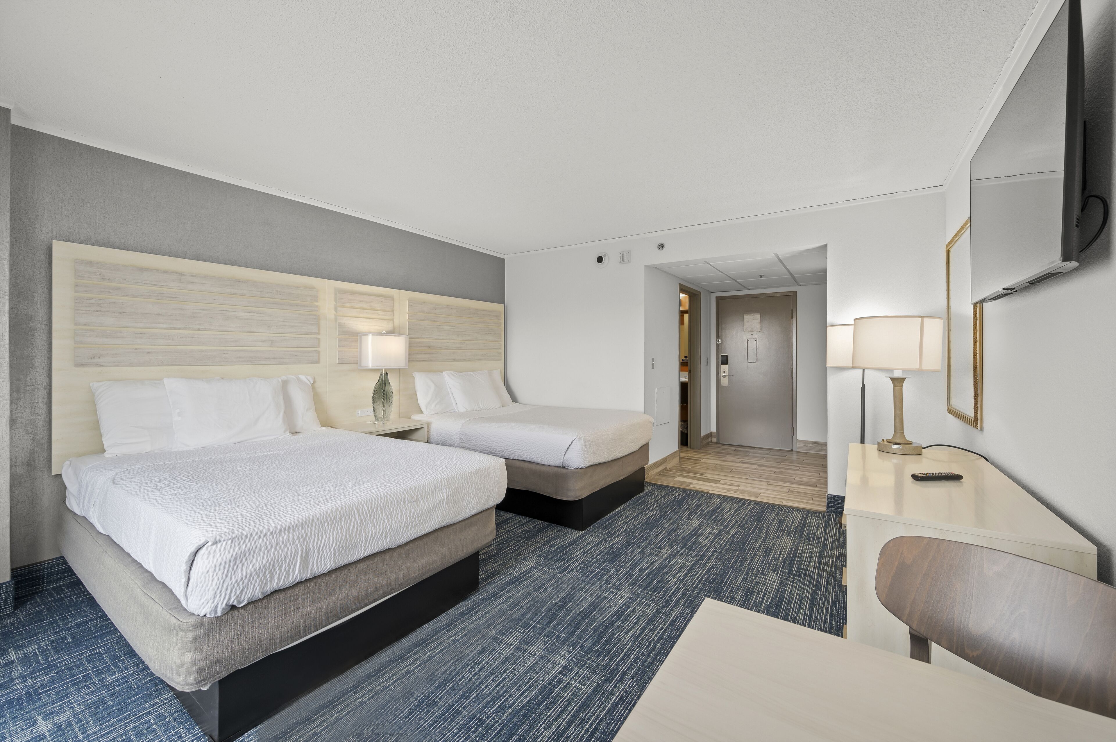 A hotel room with two beds, a desk, a chair, a TV, and a lamp.