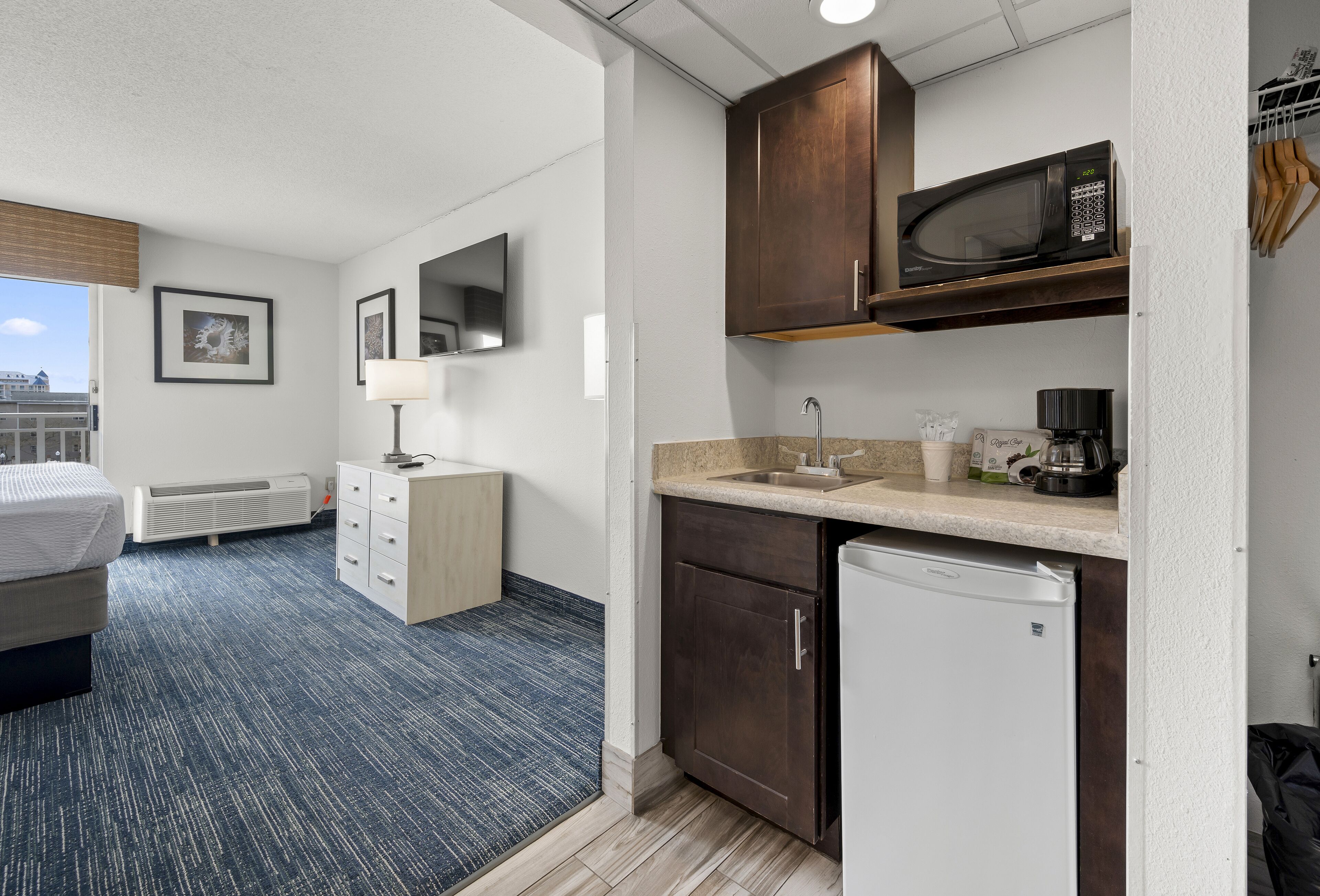 Habitación de hotel pequeña con cama, escritorio, microondas, cafetera y refrigerador.