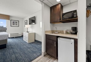 A compact hotel room with a bed, a desk, a microwave, a coffee maker, and a small refrigerator.
