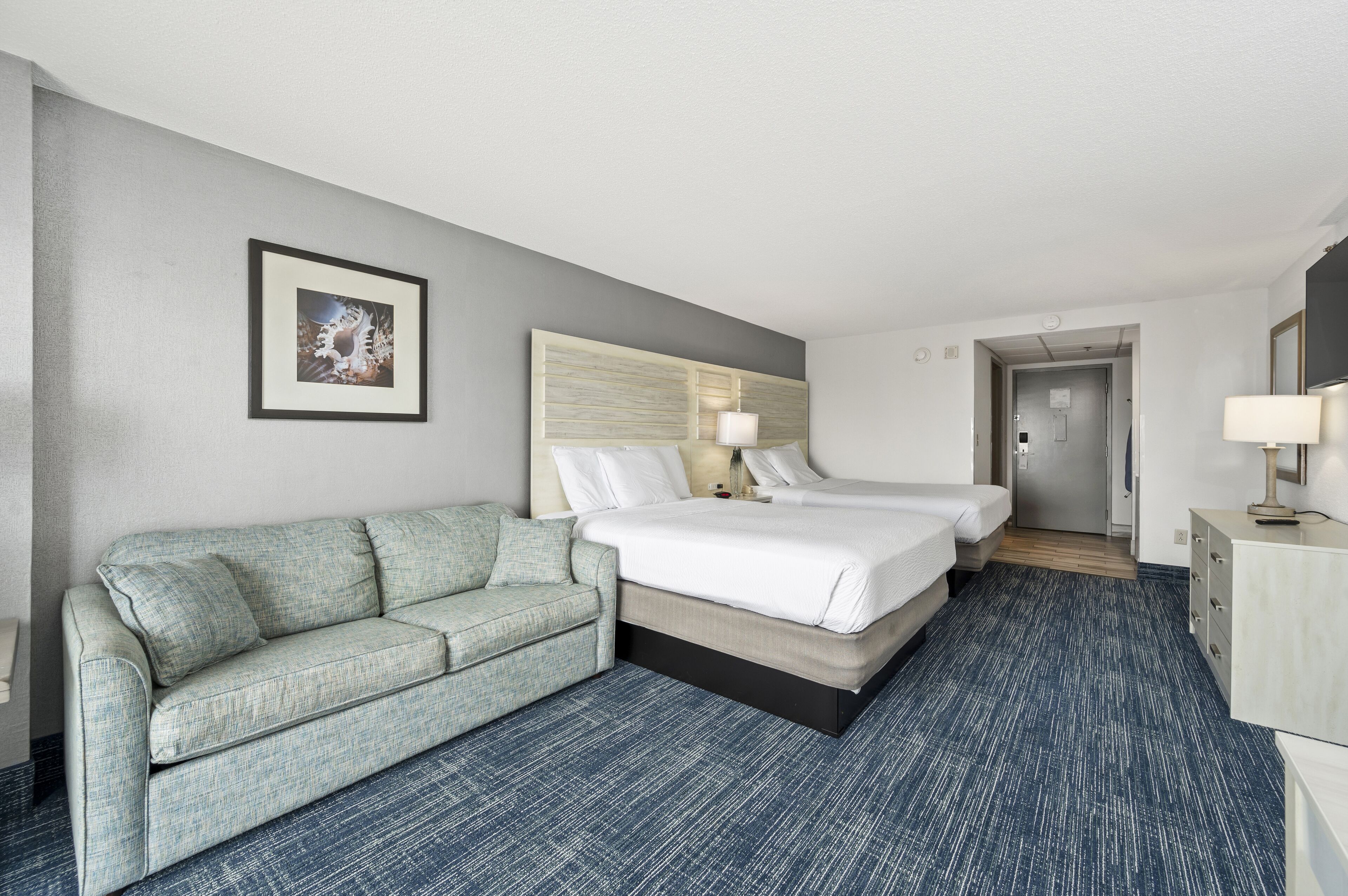 Une chambre d’hôtel avec un canapé gris, un lit avec du linge de lit blanc, une tête de lit avec une lampe et une moquette bleue.