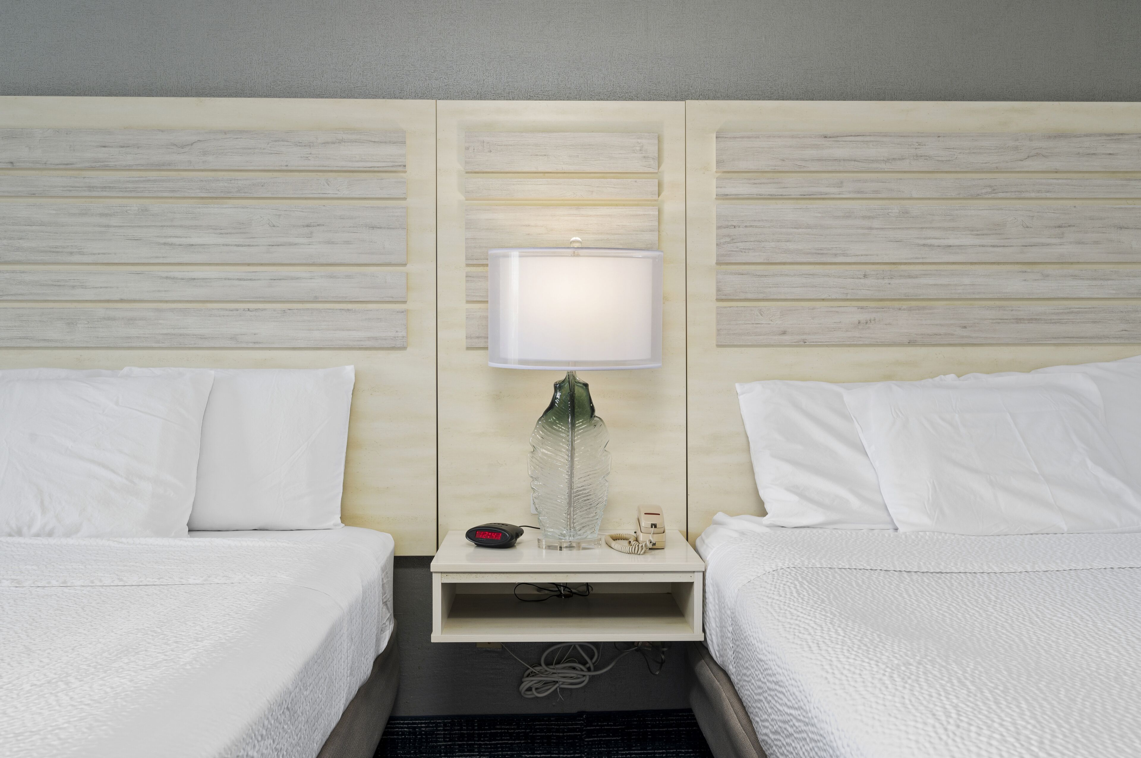 A hotel room with two beds, a nightstand with a lamp, and a small shelf with a bottle and a phone.