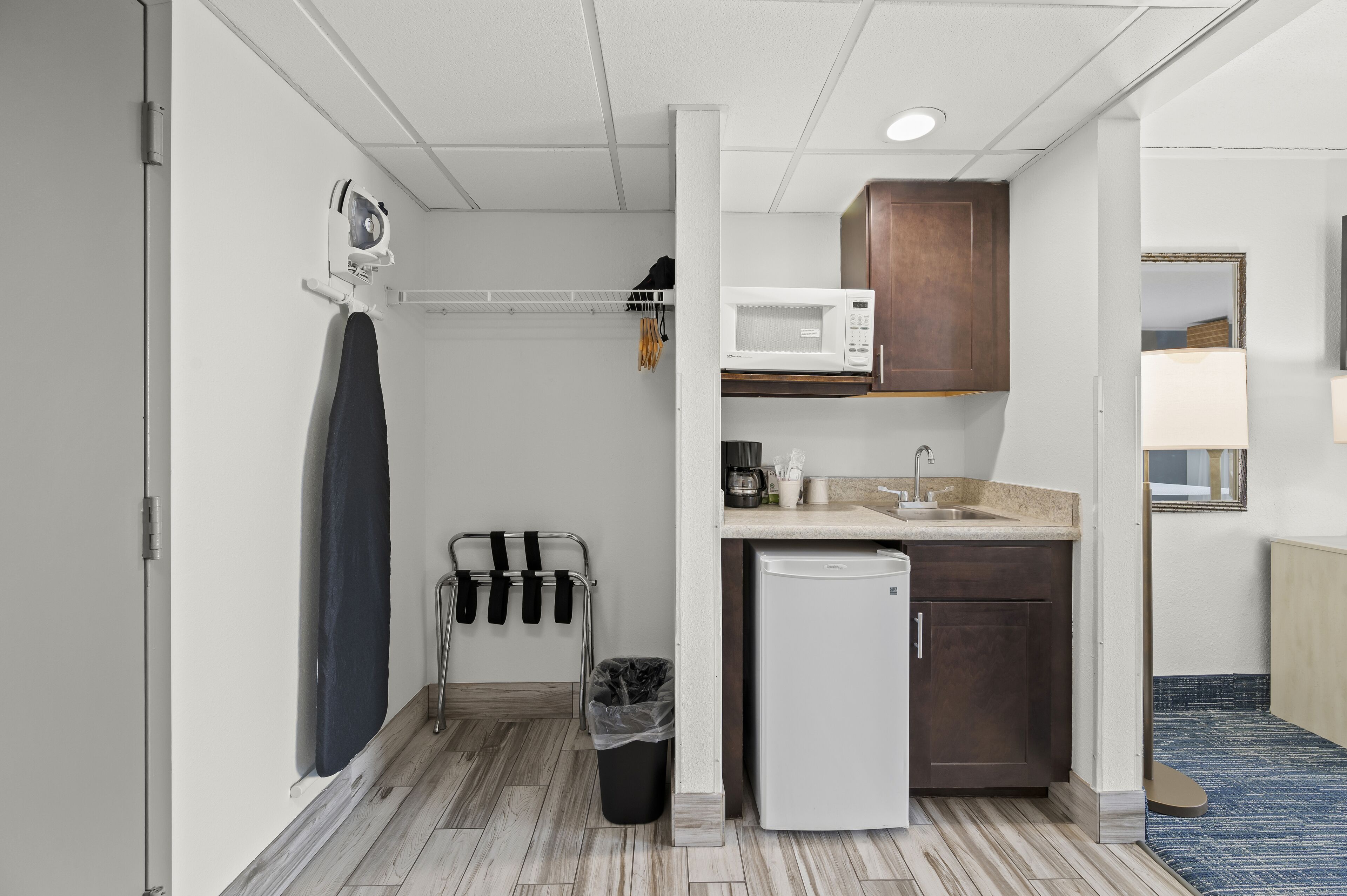 A compact kitchenette with a microwave, sink, and small refrigerator. There is a towel rack with a dark towel and a trash bin.