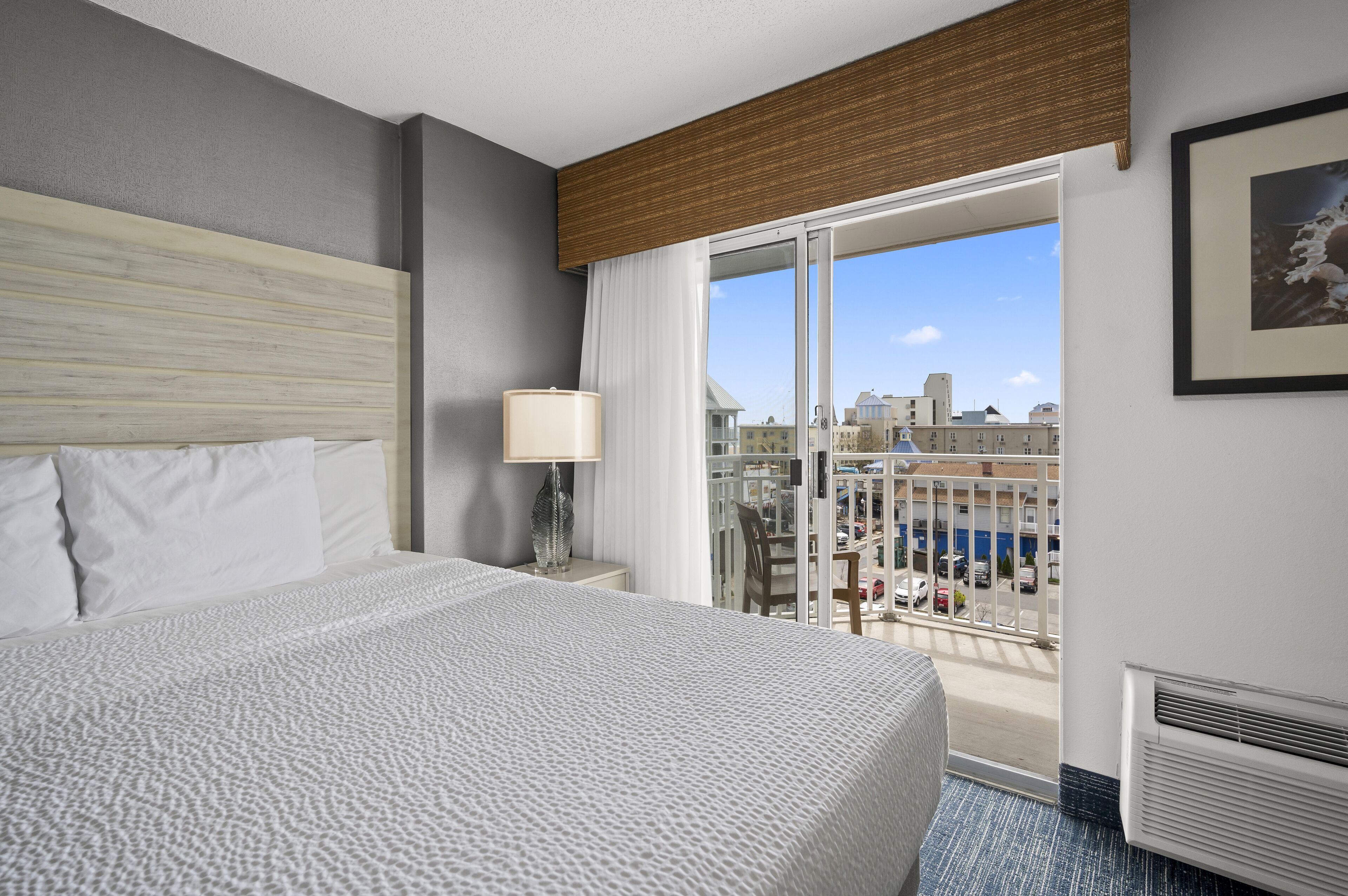 A hotel room with a large bed, a nightstand, a lamp, and a view of the city through a balcony.