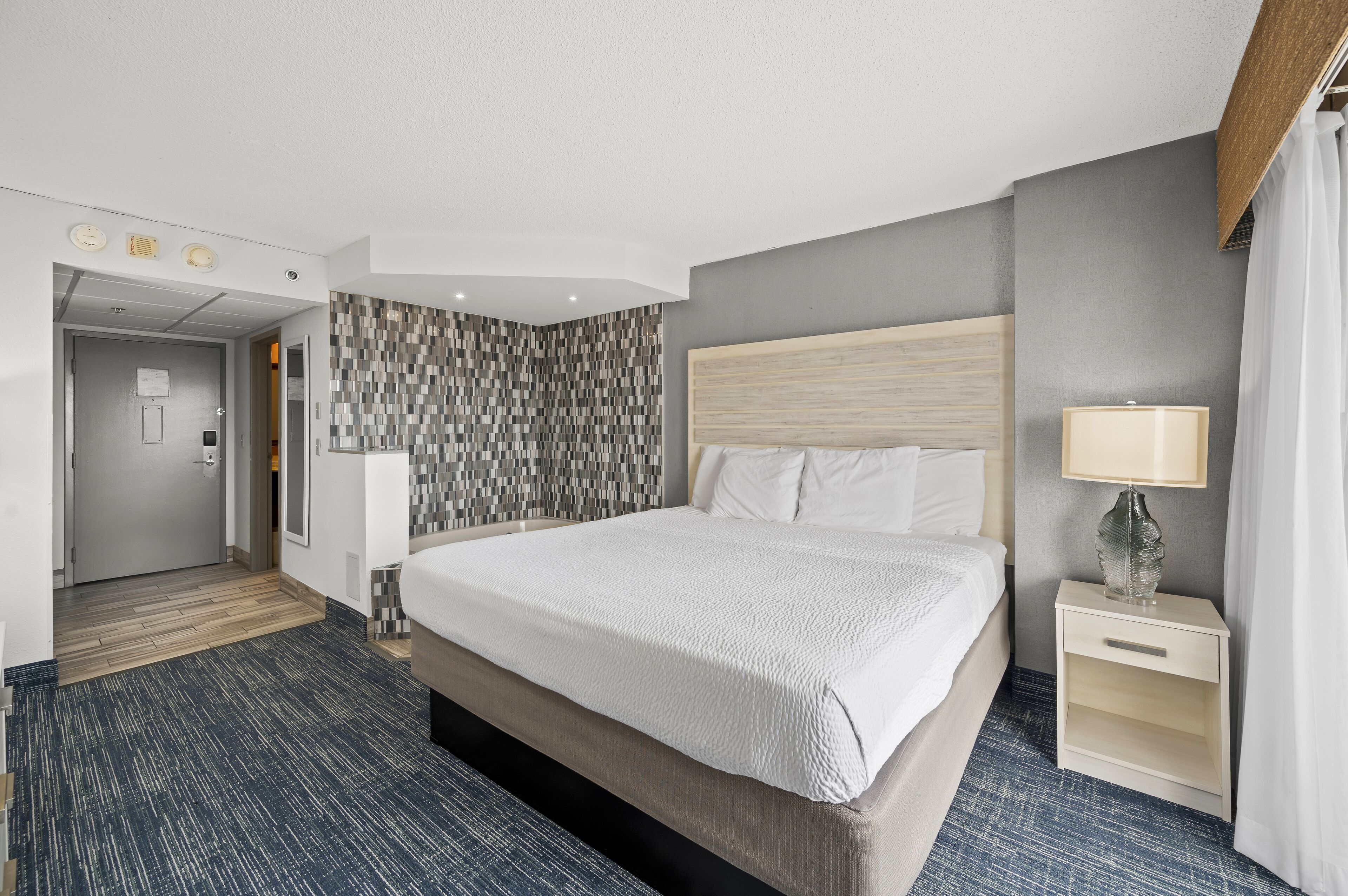A hotel room with a large bed, a bedside table with a lamp, a patterned wall, and a door leading to the hallway.