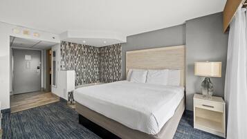A hotel room with a large bed, a bedside table with a lamp, a patterned wall, and a door leading to the hallway.