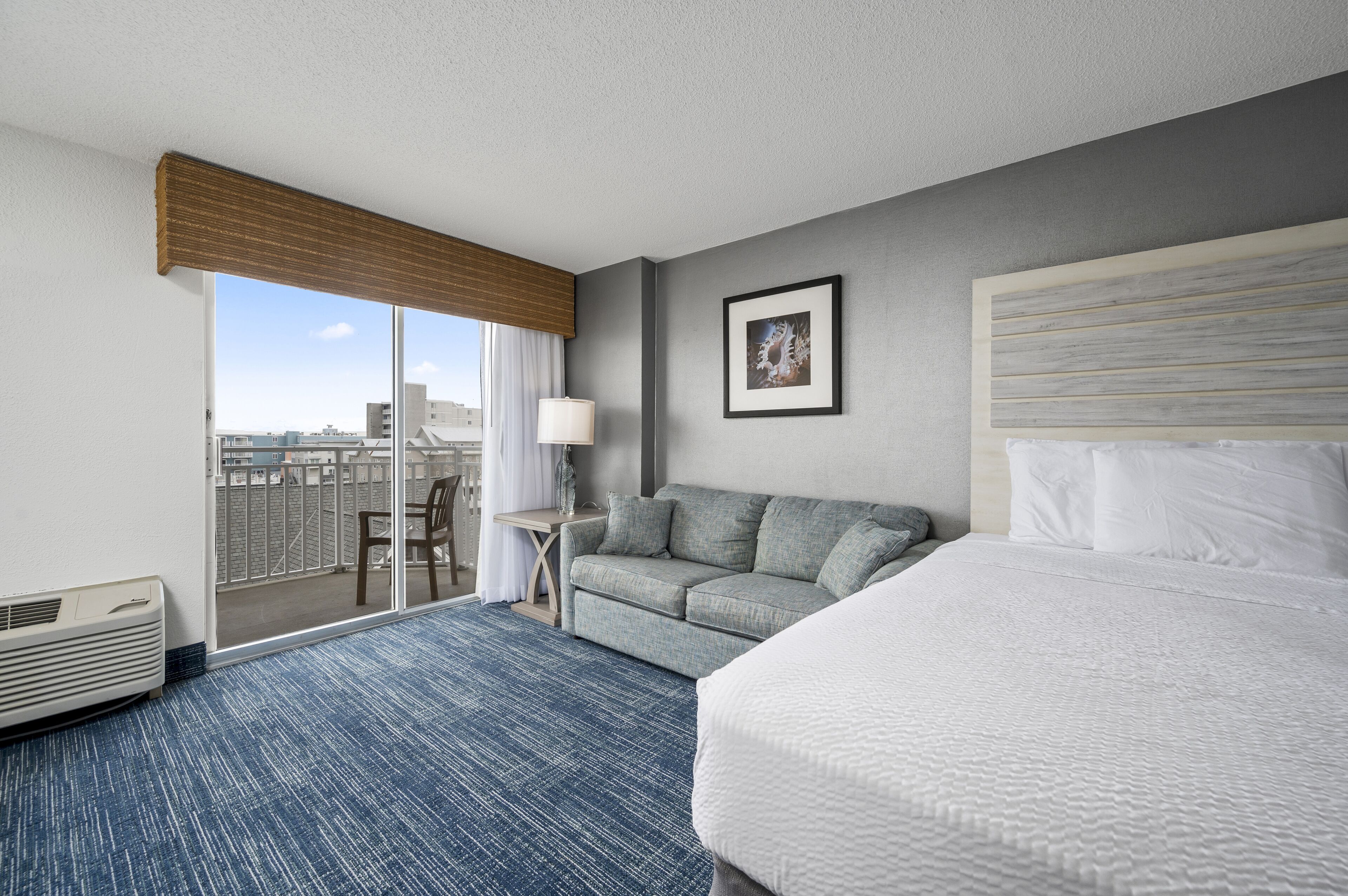 Habitación de hotel con una cama grande, un sofá, una mesita con una lámpara y vistas a la ciudad a través de una puerta corredera.
