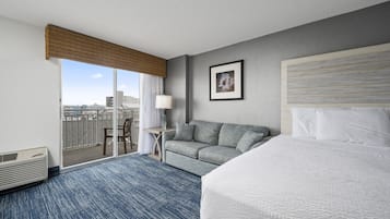 Habitación de hotel con una cama grande, un sofá, una mesita con una lámpara y vistas a la ciudad a través de una puerta corredera.