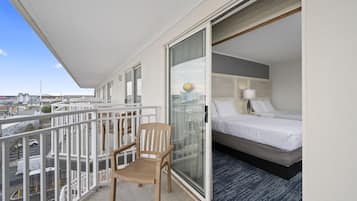 A balcony with a wooden chair, a bed with white bedding, and a city view.