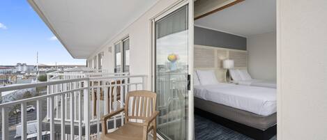A balcony with a wooden chair, a bed with white bedding, and a city view.