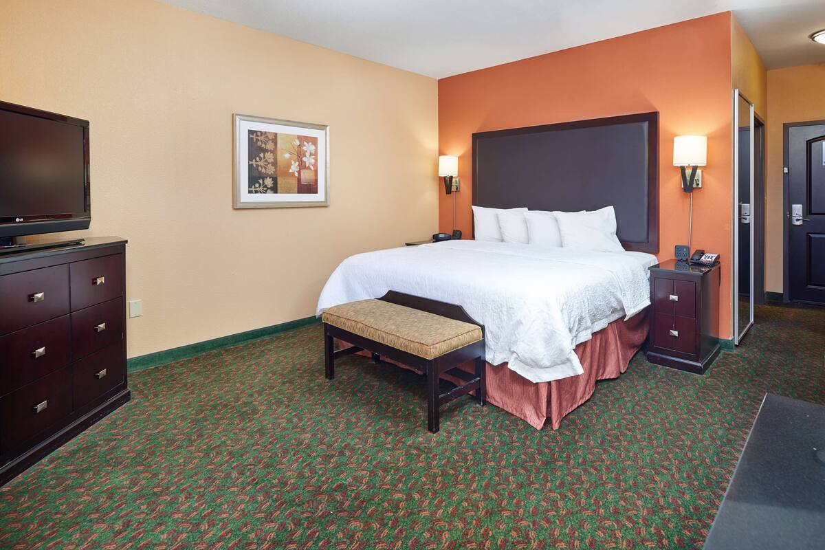 Une chambre d’hôtel avec un grand lit, un banc, une commode, une télévision et un tableau encadré au mur.