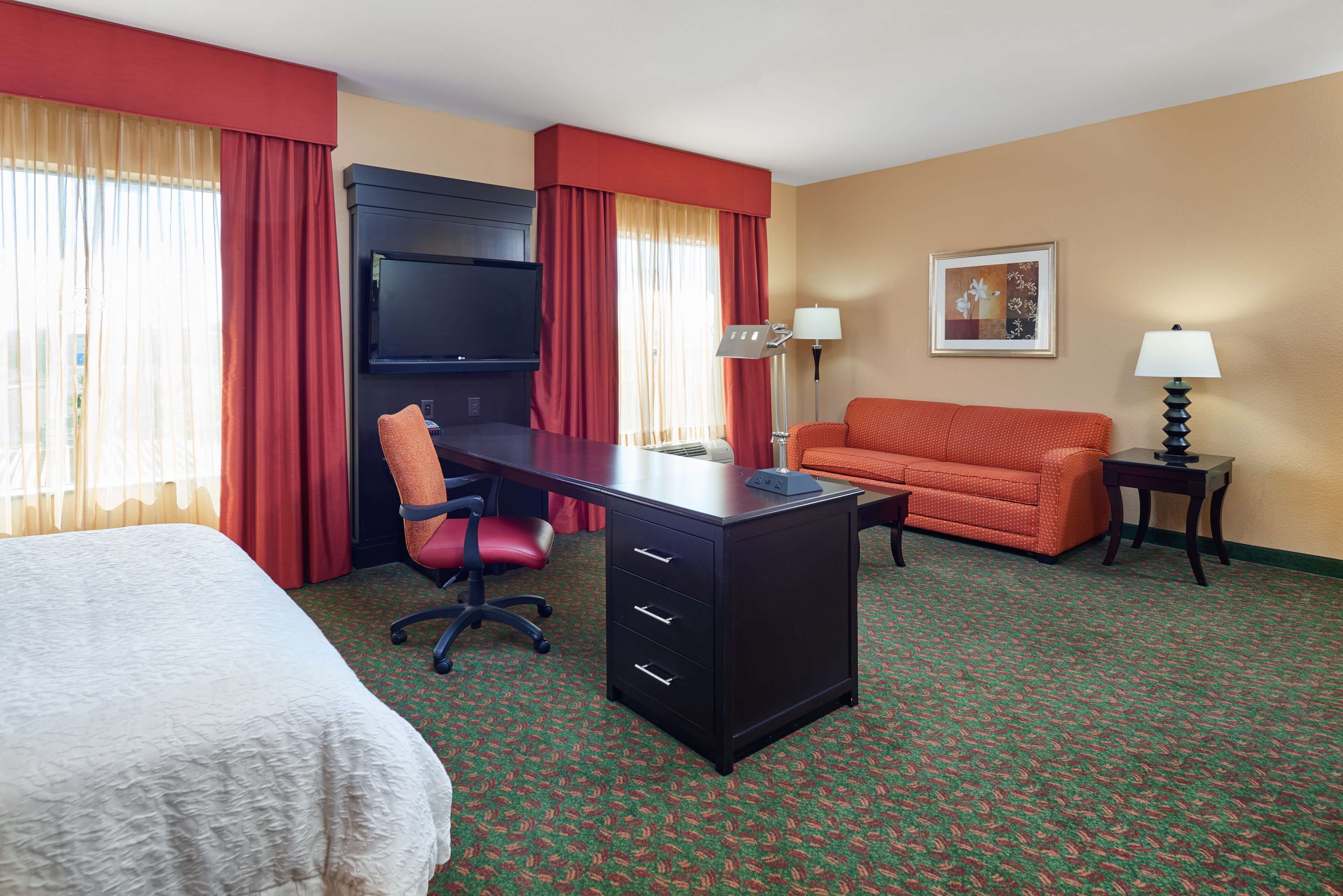A hotel room with a bed, a desk with drawers, a chair, a sofa, two lamps, and a television.