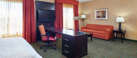 A hotel room with a bed, a desk with drawers, a chair, a sofa, two lamps, and a television.