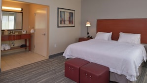 A hotel room with a bed, a bedside table with a lamp, a red suitcase, a bathroom with a sink and mirror, and a framed picture on the wall.