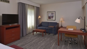 A hotel room with a TV, sofa, desk, chair, and a painting on the wall.
