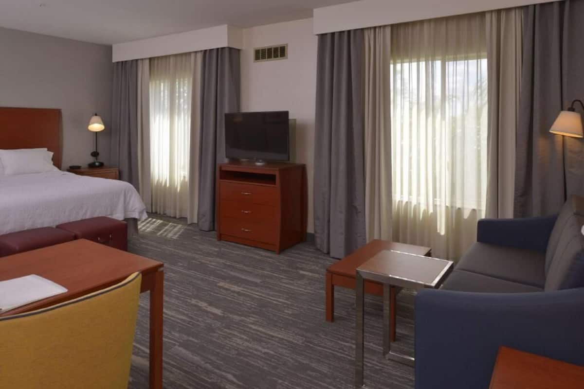 Ein Hotelzimmer mit einem Bett, einem Fernseher auf einem Holzständer, einem Schreibtisch, einer Couch und einem Fenster mit Vorhängen.