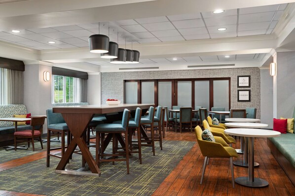 A modern hotel lobby with a central bar area, multiple seating options including bar stools and round tables, and a patterned carpet.