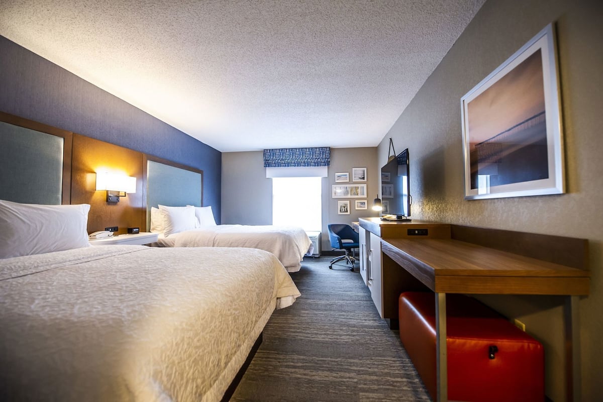 A hotel room with two beds, a desk, a chair, a TV, and a red suitcase.