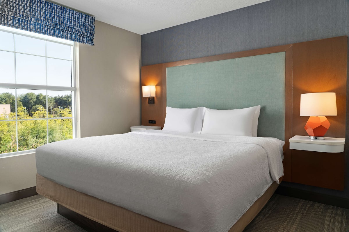 A hotel room with a large bed, two bedside tables with lamps, a window with a view of trees, and a headboard with a textured panel.
