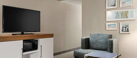 Una habitación de hotel moderna con una televisión de pantalla plana, microondas, una pequeña mesa, un sillón con cojín y cuadros en la pared.