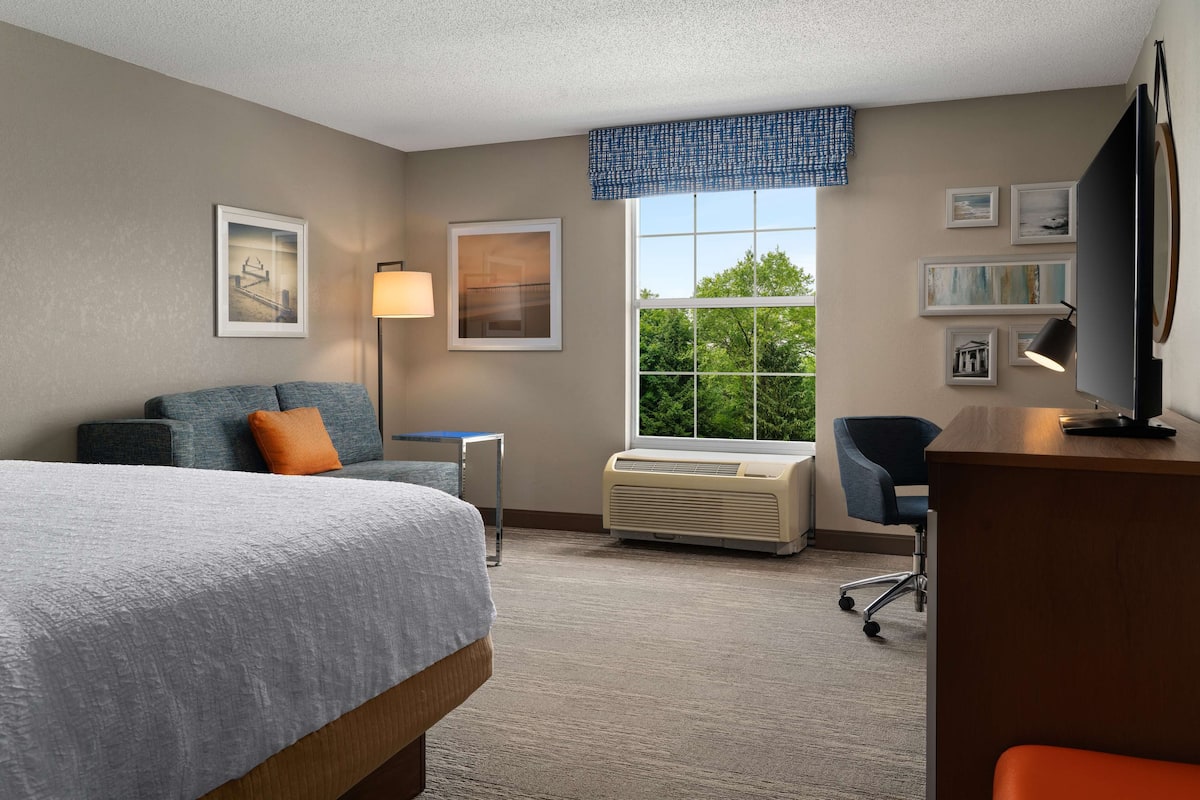 A hotel room with a bed, a desk, a chair, a lamp, an air conditioner, and framed pictures on the wall.