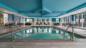 Una amplia piscina interior con agua cristalina y azul, rodeada de sillas de descanso y mesas, y un techo azul.