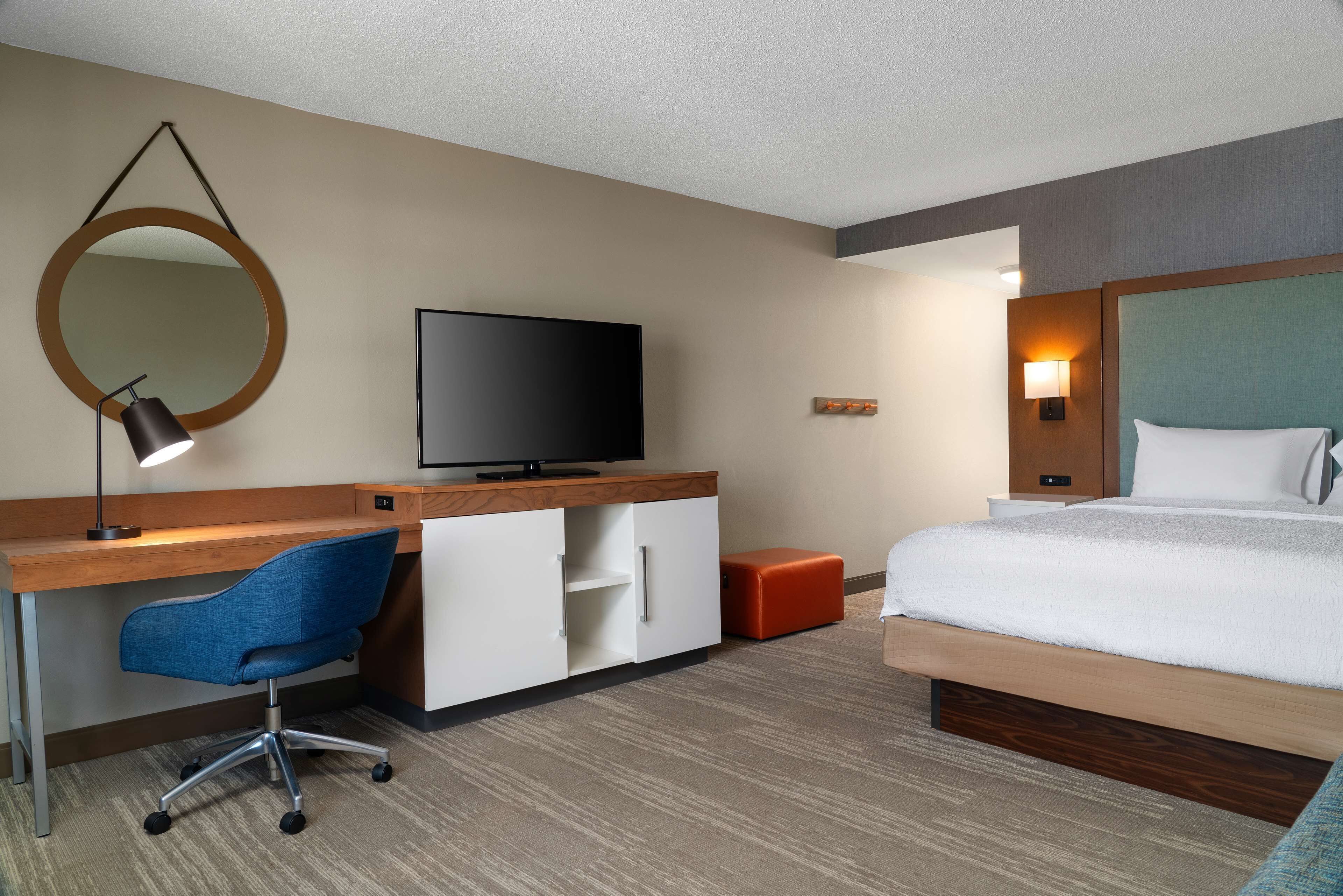 Een hotelkamer met een houten bureau, een blauwe bureaustoel, een televisie, een bed met wit beddengoed, een ronde spiegel en een klein oranje poef.