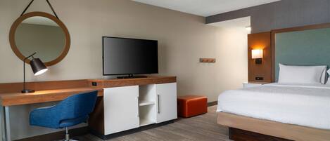 Een hotelkamer met een houten bureau, een blauwe bureaustoel, een televisie, een bed met wit beddengoed, een ronde spiegel en een klein oranje poef.