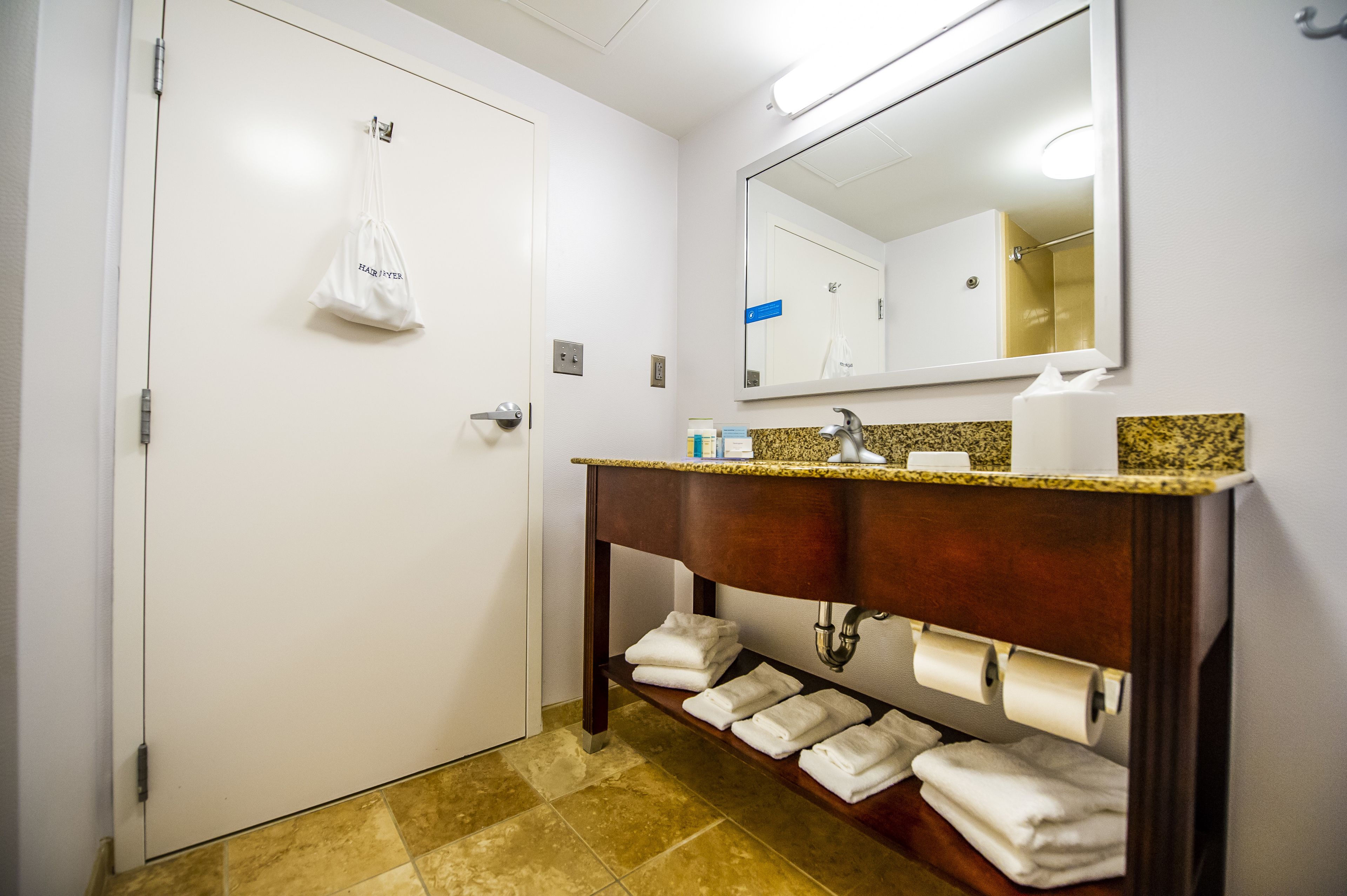 Un baño de hotel con una puerta blanca, un espejo, un lavamanos con encimera de granito y un estante con toallas dobladas.