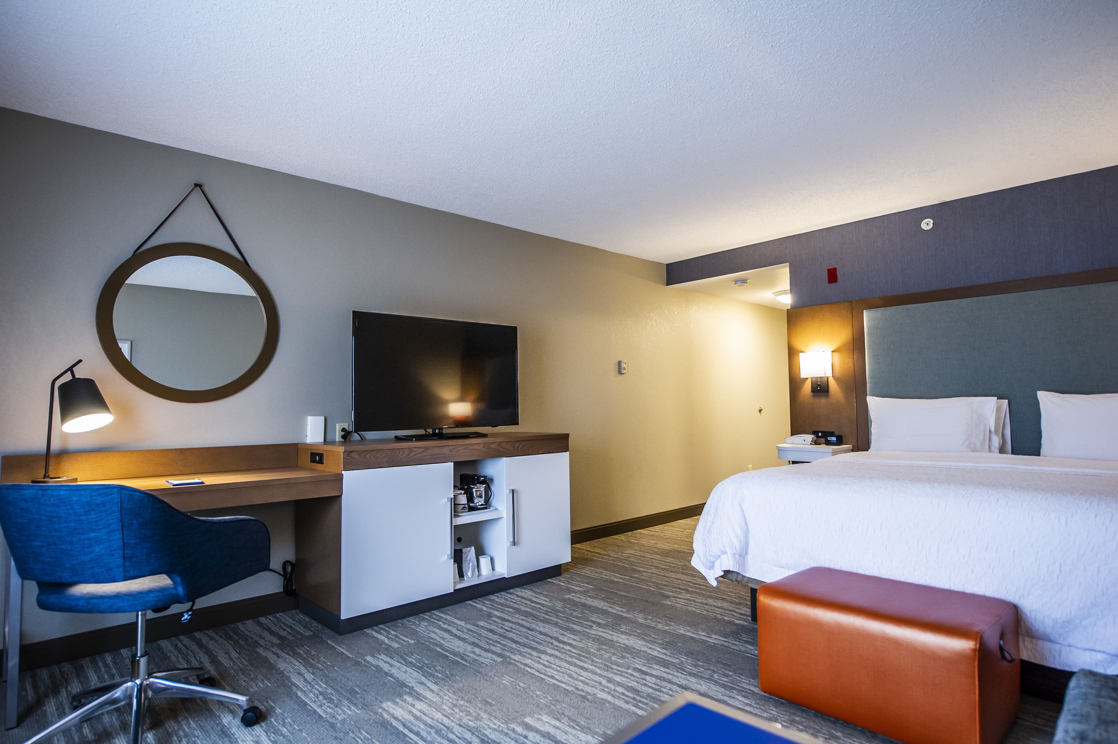 Ett hotellrum med en säng, ett skrivbord med en stol, en tv, en spegel och ett litet bord med en lampa.
