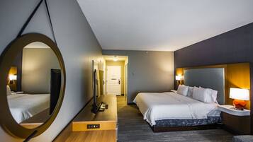 Habitación de hotel con un espejo grande, una cama, un escritorio con computadora y una mesita de noche con lámpara.
