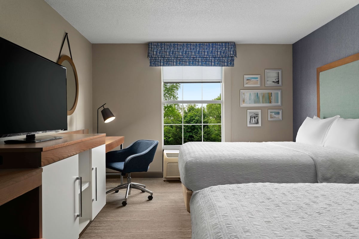 A hotel room with a large bed, a desk with a flat-screen TV, a chair, a window with a view of trees, and several framed pictures on the wall.