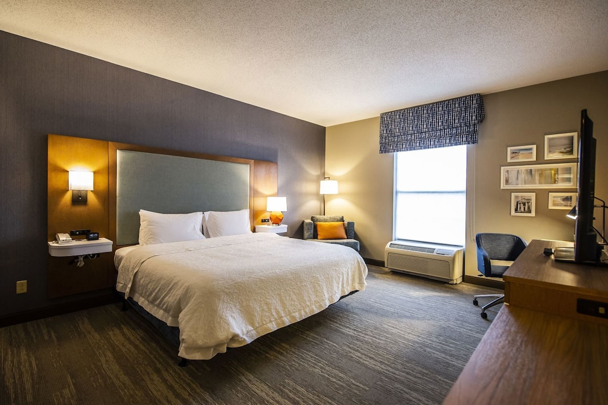 A hotel room with a bed, bedside tables, a desk, a chair, a window with blinds, and wall-mounted artwork.