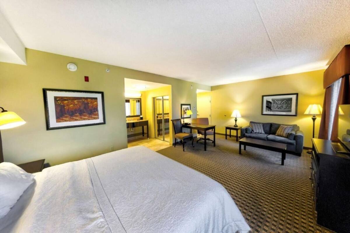 A hotel room with a large bed, a sofa, a desk, and a chair. There is a mirror, a lamp, and a picture on the wall.
