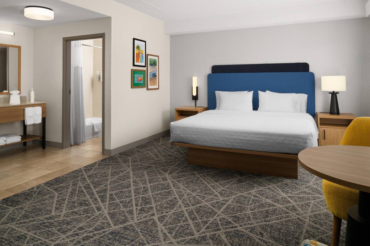 A hotel room with a bed, bedside tables, a bathroom, a wooden chair, and a patterned rug.