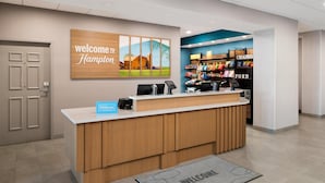 A hotel reception desk with a 'Welcome to Hampton' sign, a digital kiosk, and a snack bar in the background. - Hampton Inn & Suites Chadds Ford (Glen Mills)