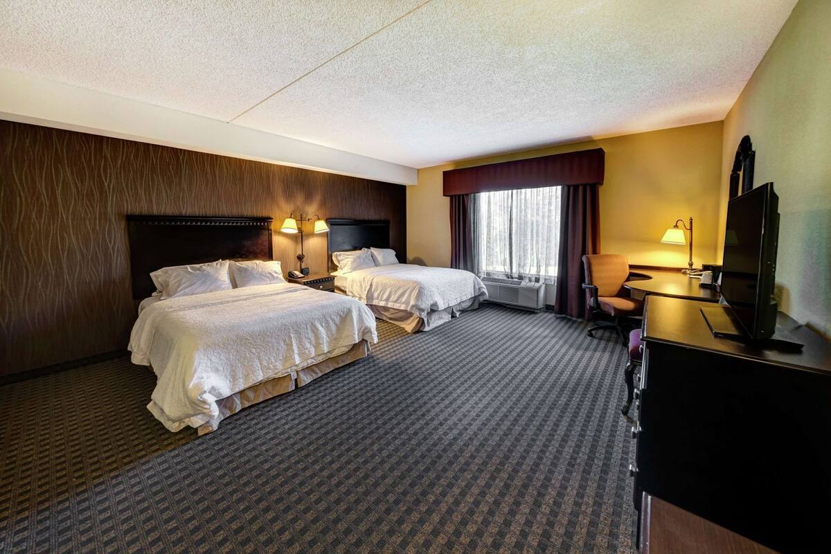 A hotel room with two beds, a desk with a TV, a chair, and a window with curtains.