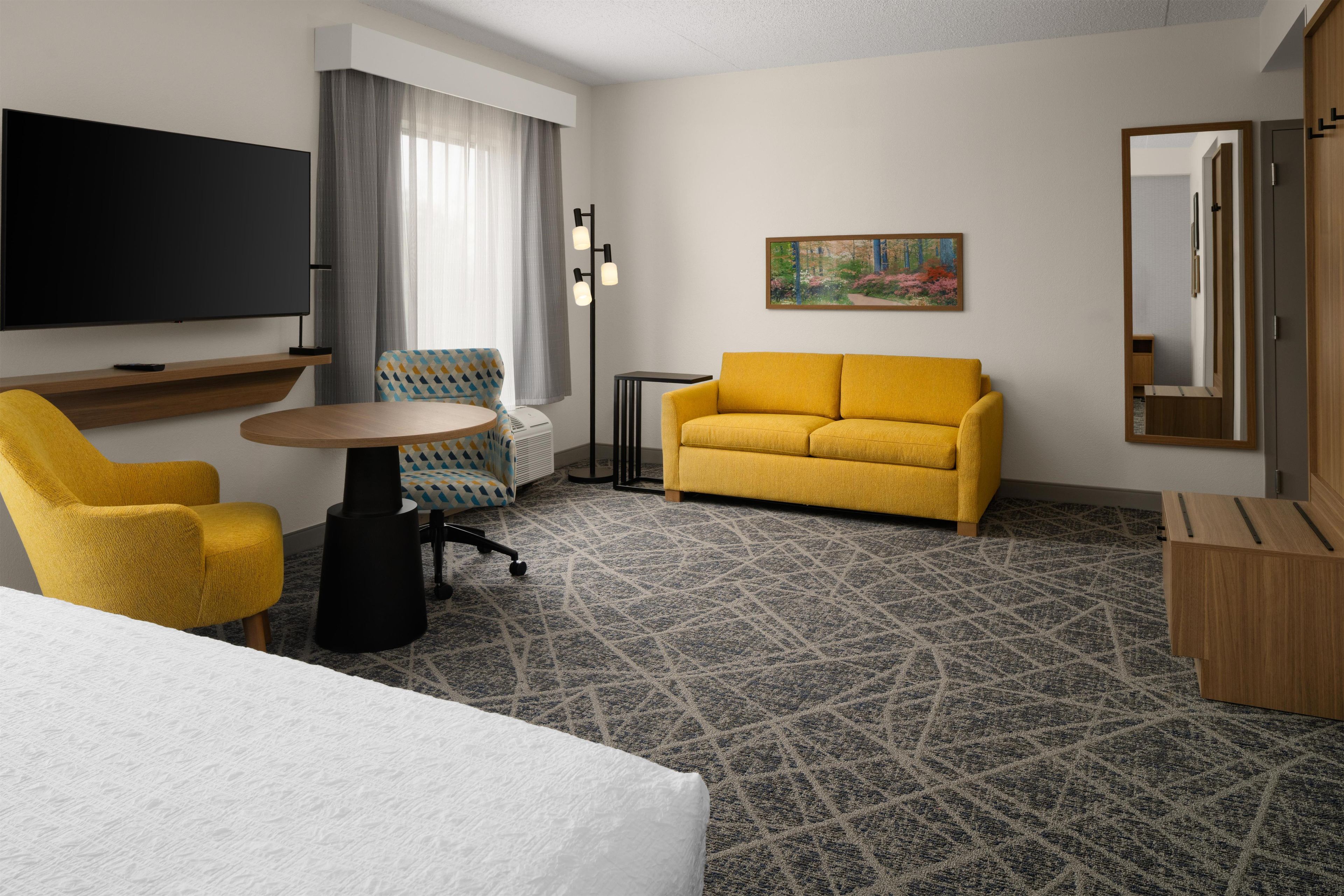 Habitación de hotel con cama, sofá amarillo, una mesita, televisor, un cuadro y un espejo.