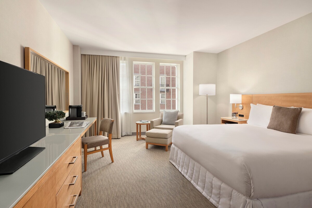 Une chambre d’hôtel comprenant un lit, un bureau avec une télévision, une chaise, une petite table et une fenêtre avec des rideaux.
