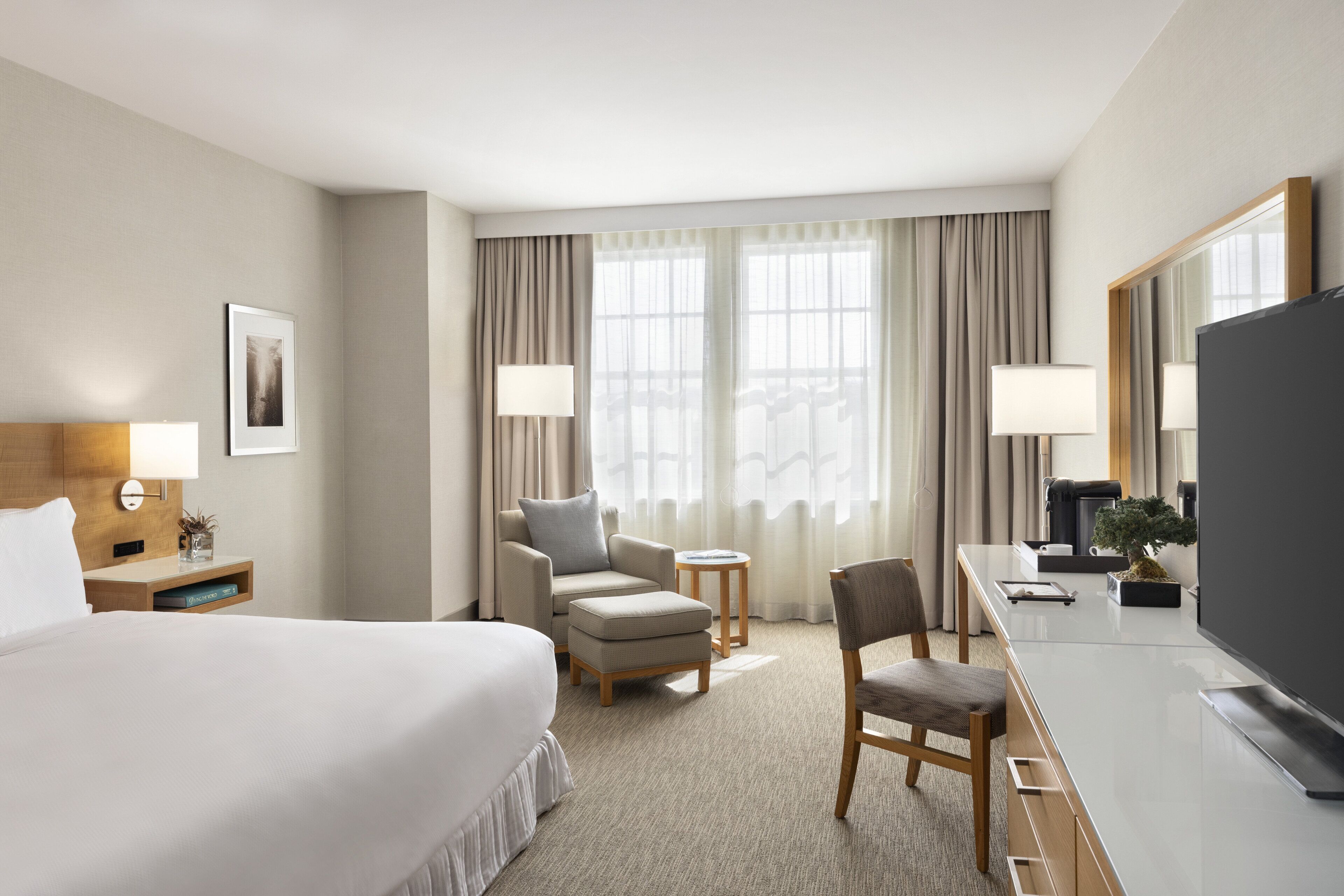 A hotel room with a large bed, a desk with a flat-screen TV, a chair, a small table, and a window with curtains.