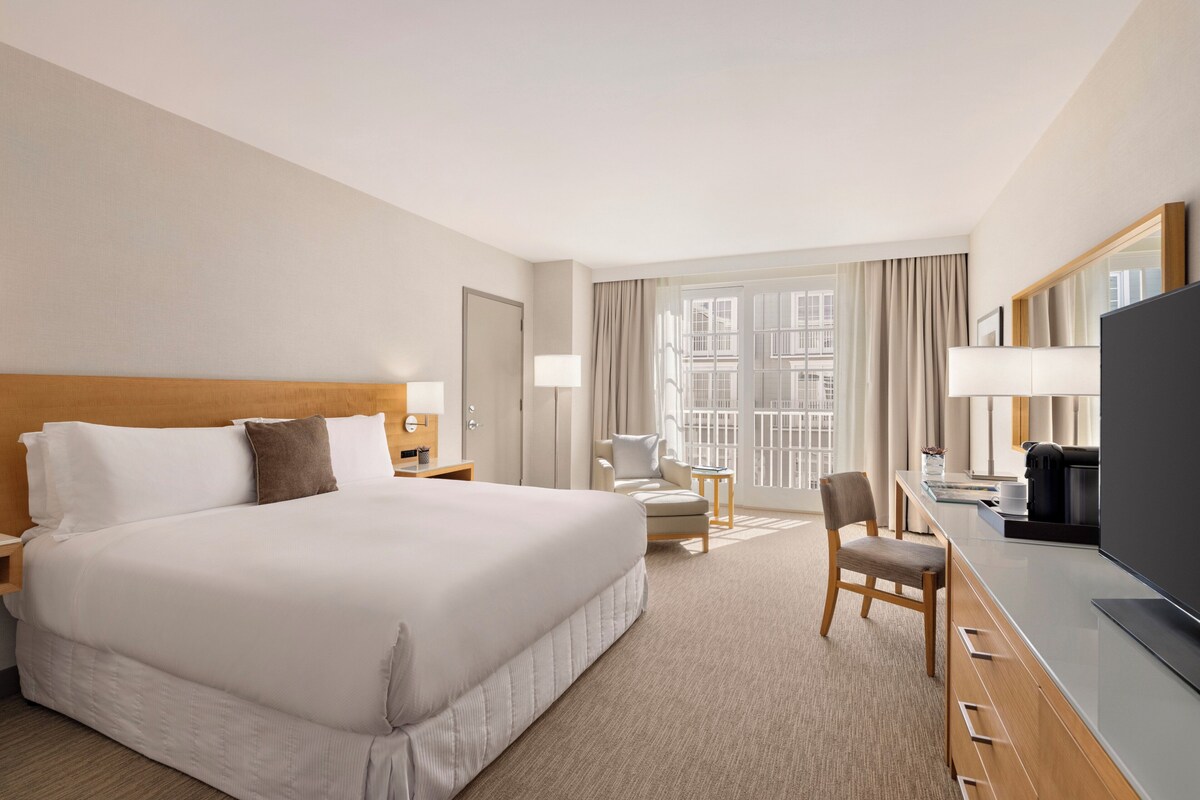 Une chambre d’hôtel avec un grand lit, un bureau avec une chaise, une télévision et une fenêtre avec des rideaux.