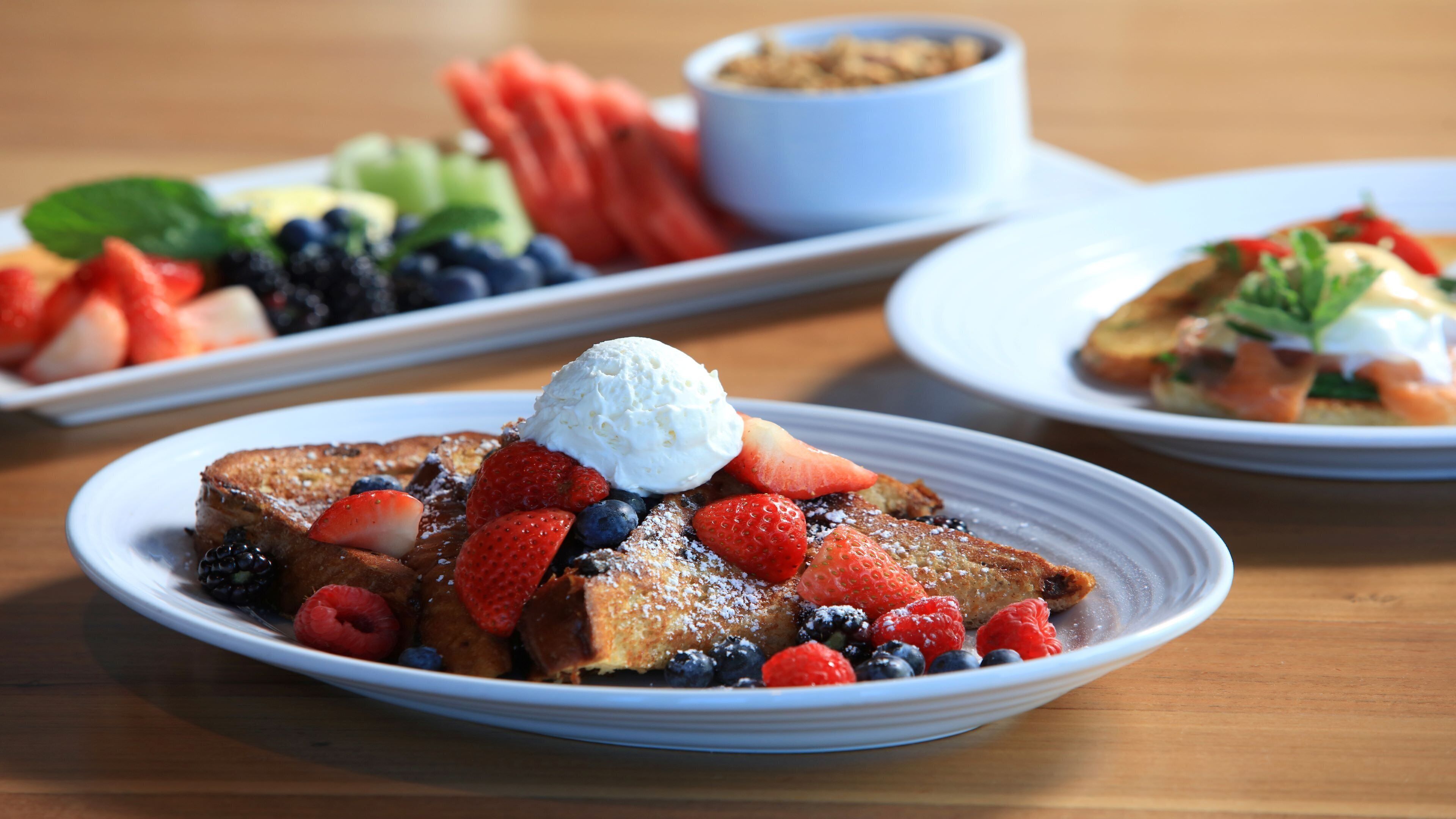 A breakfast setting with French toast topped with strawberries, blueberries, raspberries, and whipped cream, served on white plates.