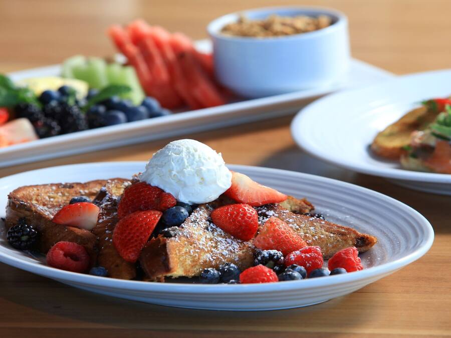 En morgenmadsanretning med French toast pyntet med jordbær, blåbær, hindbær og flødeskum, serveret på hvide tallerkener.