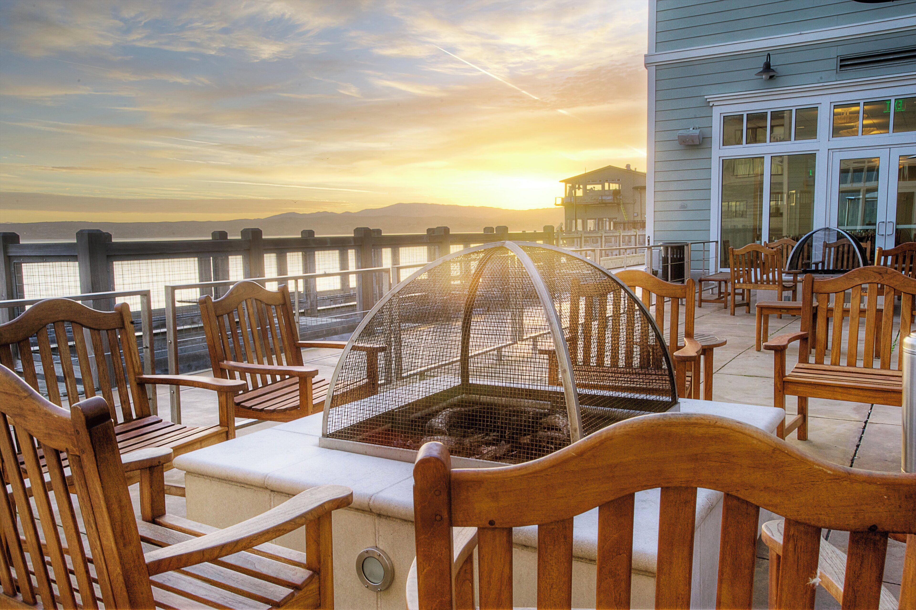 Una terraza con sillas de madera y cerramiento de vidrio, con vista a un cuerpo de agua y un atardecer de fondo.