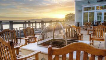 Una terraza con sillas de madera y cerramiento de vidrio, con vista a un cuerpo de agua y un atardecer de fondo.