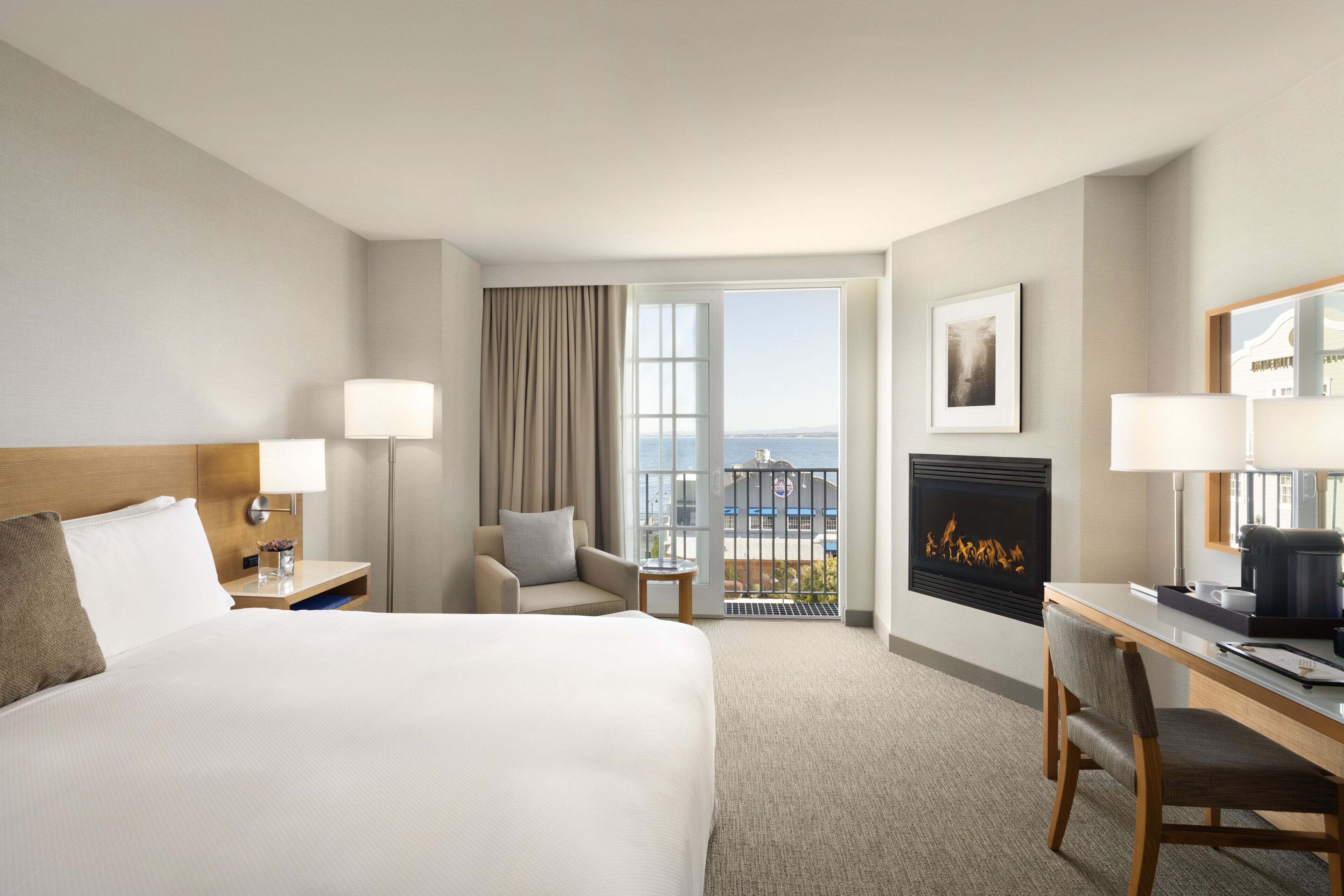 A hotel room with a large bed, a desk, a chair, a fireplace, a balcony with a view, and a nightstand with a lamp.
