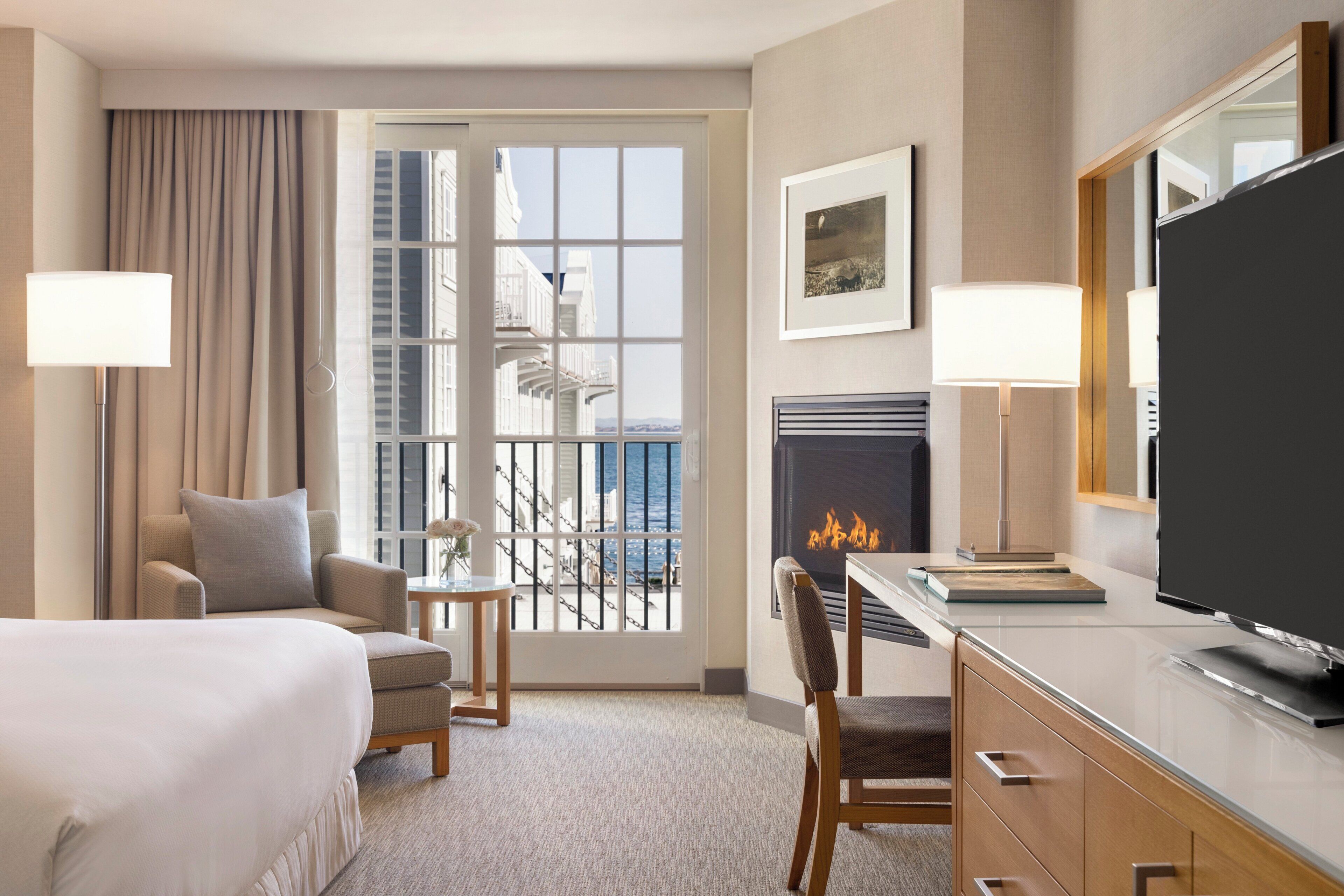 A hotel room with a bed, a desk, a chair, a television, a fireplace, and a view of the sea through a balcony door.
