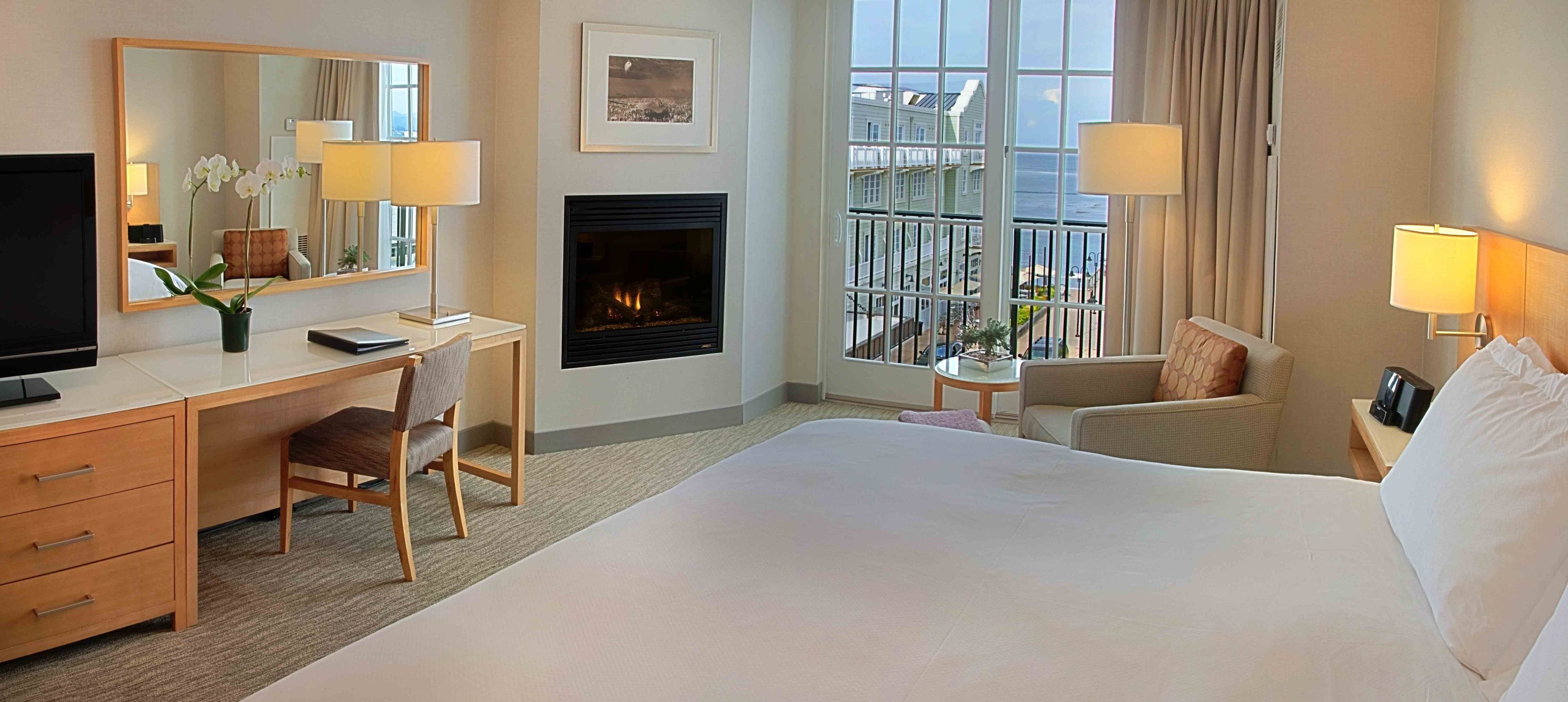 A hotel room with a large bed, a desk with a chair, a TV, a fireplace, and a balcony with a view of the city.