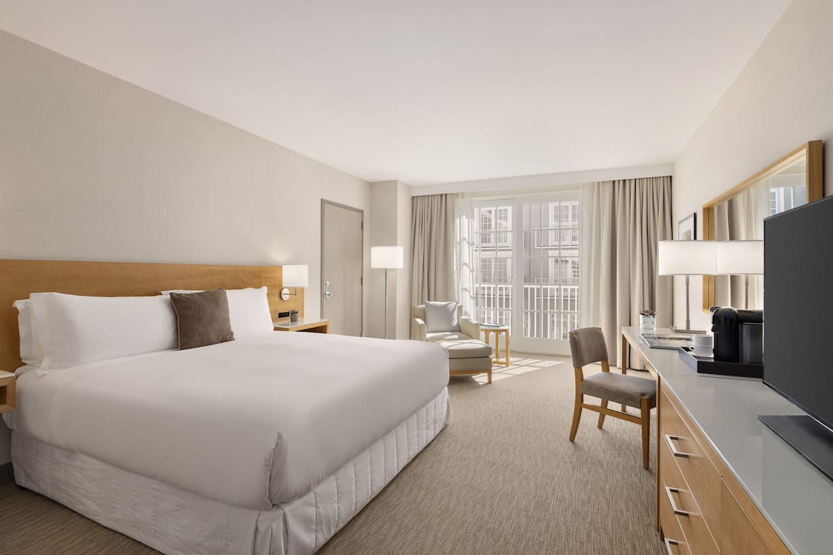 Quarto de hotel com uma cama grande, uma mesa de trabalho com televisão, uma cadeira e uma janela com cortinas.