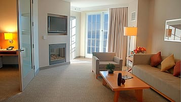 Habitación de hotel con sofá, mesa de centro, televisor, chimenea y un ventanal con cortinas.